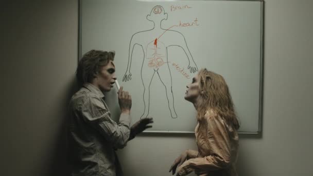 Tiro medio del hombre zombi apuntando a la imagen de la persona dibujada en la pizarra blanca y explicando las partes del cuerpo humano a la mujer zombi - Metraje, vídeo