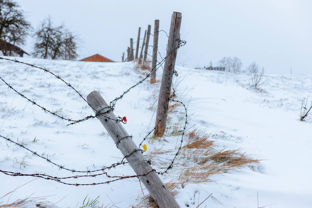 Barbed wire fence with fallen pillars between two grazing grounds in winter snowy Bavarian mountain landscape. - Photo, Image
