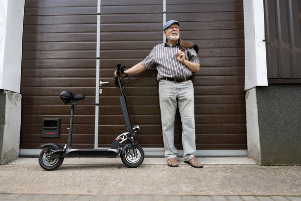 Positiivinen, iäkäs mies seisoo oven lähellä ja pitää ekoskootteria pyörän takana. Ikääntyneiden terveellinen elämäntapa. - Valokuva, kuva