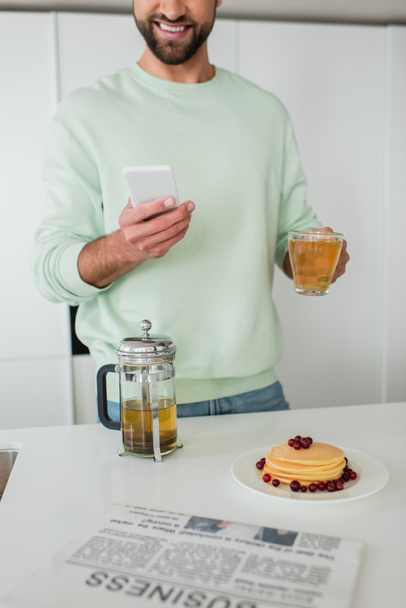 vista parcial del hombre sonriente sosteniendo teléfono inteligente y té verde cerca de panqueques en la cocina - Foto, imagen
