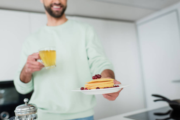 cropped view of happy blurred man holding green tea and plate with pancakes  - Photo, Image