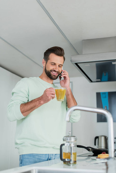 веселый человек с чашкой зеленого чая разговаривает по мобильному телефону на кухне - Фото, изображение