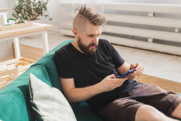 Homme détendu utilisant un smartphone assis sur un canapé vert dans le salon à la maison - Photo, image