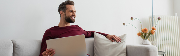 smiling man looking away while sitting on couch with laptop, banner - Foto, Imagen