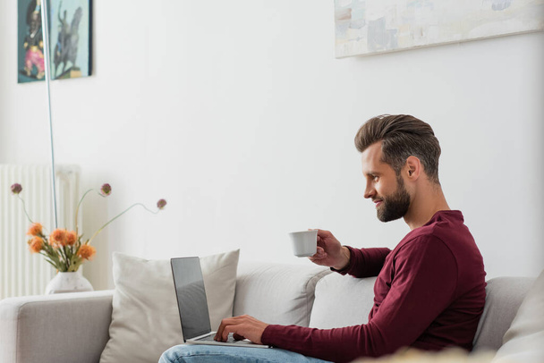 uomo in possesso di una tazza di caffè mentre seduto sul divano con computer portatile - Foto, immagini