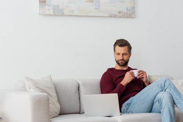 man with cup of coffee sitting on sofa near laptop - Photo, Image