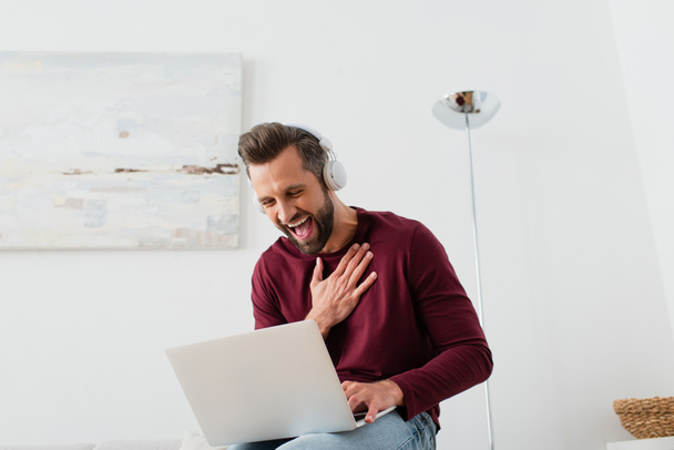Kulaklıklı neşeli adam evde dizüstü bilgisayarın yanında gülüyor. - Fotoğraf, Görsel