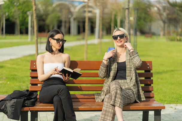 Two businesswomen are sitiing on the bench while smiling and checking the note book - Foto, Bild