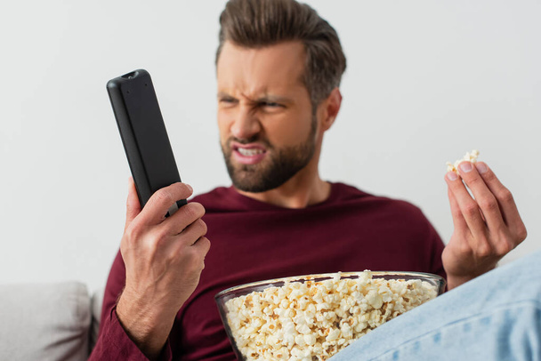 displeased man with popcorn looking at tv remote controller, blurred background - Fotografie, Obrázek