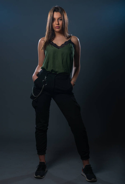 Портрет сексуальной молодой женщины-модели, стоящей в студии и одетой в темно-зеленый топ и черные брюки - Фото, изображение