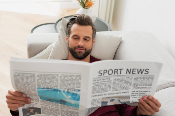 tevreden man leest sport nieuws op de bank thuis - Foto, afbeelding