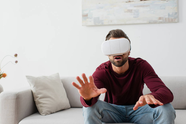 astonished man gesturing while sitting on couch in vr headset - Photo, Image