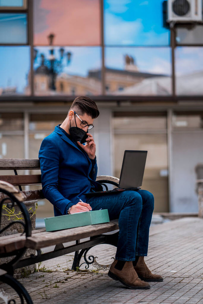 Fashionable businessman using laptop while sitting on wooden bench and talking on mobile phone - Photo, Image