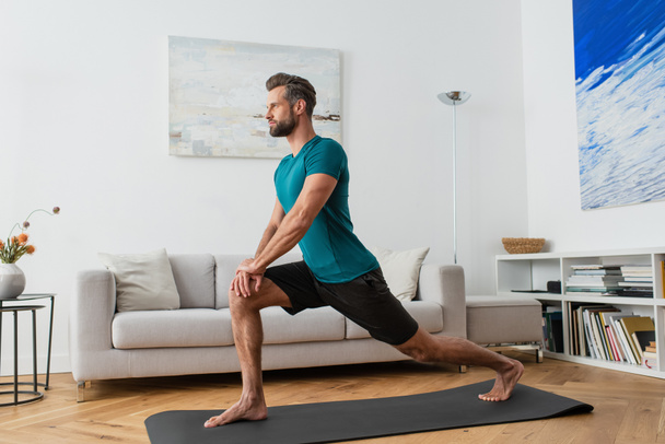 sportive man practicing crescent lunge pose on yoga mat at home - Фото, зображення