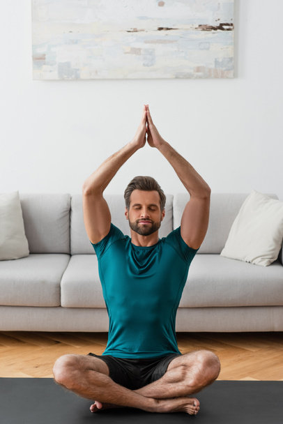 man with praying hands and closed eyes meditating in lotus pose at home - Fotografie, Obrázek