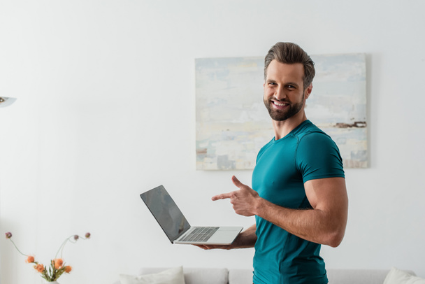happy man pointing with finger at laptop while smiling at camera - Foto, Bild