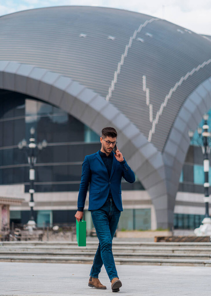 Happy young man wearing business suit while talking on mobile phone and walking along a modern corporate building - Foto, immagini