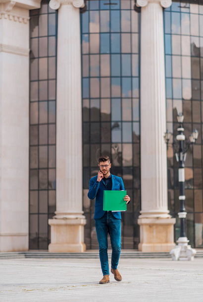 Selbstbewusster Geschäftsmann hält grünen Ordner in der Innenstadt und telefoniert - Foto, Bild