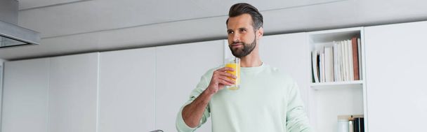 joyful man looking away while holding glass of orange juice in kitchen, banner - Photo, image