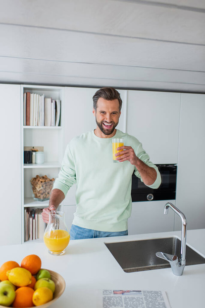 laughing man holding glass of fresh orange juice in kitchen - Fotó, kép