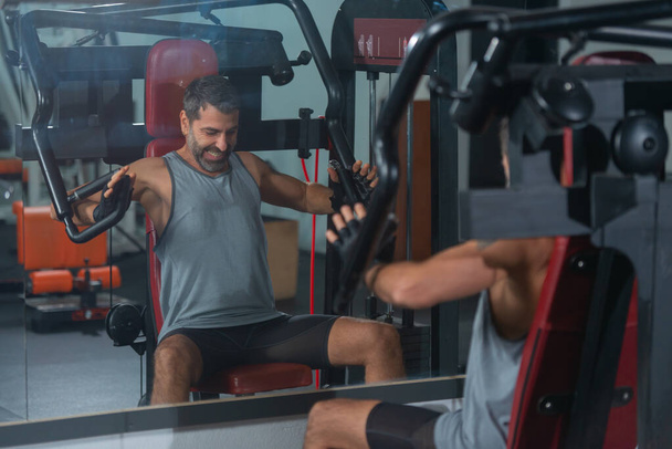 Изображение в зеркале сильного мускулистого человека тренирующего мышцы спины с гирями на тренажере в тренажерном зале - Фото, изображение