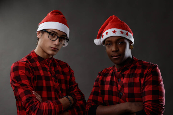 Двоє серйозних вдумливих молодих чоловіків у святкових червоних різдвяних капелюхах стоять поруч, дивлячись на камеру зі складеними руками на сірому студійному фоні
 - Фото, зображення