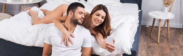 Smiling woman in lingerie taking selfie near boyfriend in bed, banner  - Photo, Image