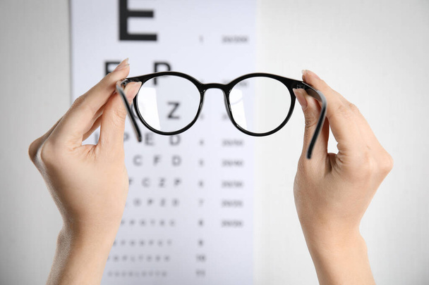 Woman holding glasses against eye chart on light background, closeup. Ophthalmologist prescription - Fotoğraf, Görsel