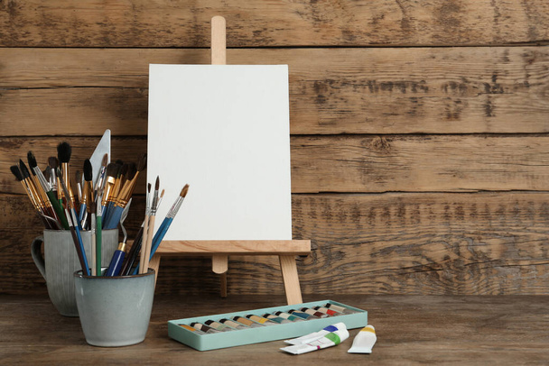 Easel with blank canvas, paints and brushes on wooden table. Space for text - Фото, зображення