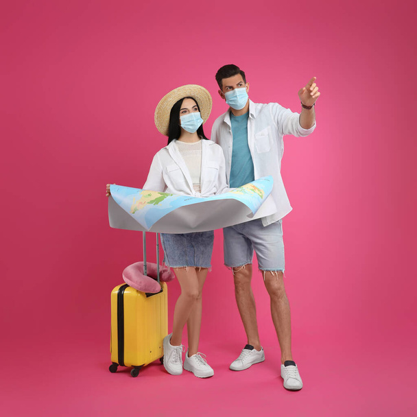 Couple of tourists in medical masks with map and suitcase on pink background. Travelling during coronavirus pandemic - Foto, Imagem
