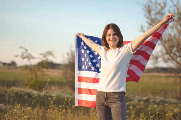 Egy gyönyörű fiatal nő tartja Amerika nemzeti zászlaját, miközben egy mezőn áll naplementekor. Július 4., Függetlenség Napja. Amerikai szabadság fogalma. - Fotó, kép