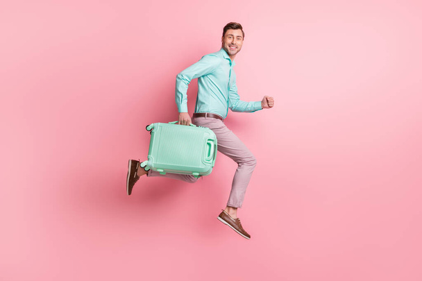 Full size profile side photo of brunette man hold luggage trip run jump wear teal shirt pink pants isolated on pastel color background - Foto, immagini