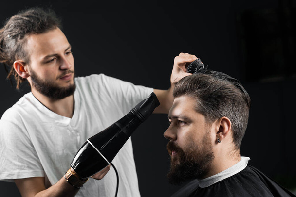 床屋はファッショナブルな髪型の後にハンサムな髭の男の髪を乾燥させます。検疫期間中の美容師の仕事コロナウイルスcovid-19 - 写真・画像