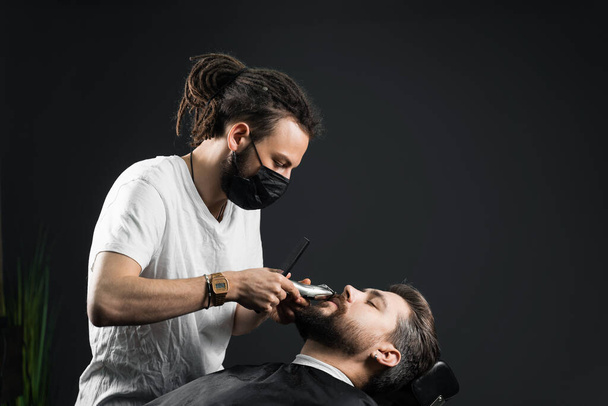 El bigote se afeita en la peluquería. Peluquero con rastas en máscara médica negra recortar la barba del hombre guapo en la cuarentena coronavirus covid-19 - Foto, imagen