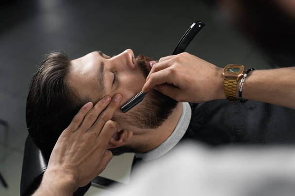 Прямая бритва стрижет бороду в парикмахерской. Парикмахер делает прическу красивому мужчине - Фото, изображение