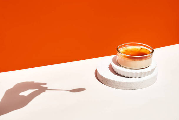 キャラメリゼした黒糖オレンジの背景を持つクリームブリュレフレンチデザート。おいしいカタログクリームの2つの部分。表彰台と影を持つ創造的な食品。コピースペース。選択的焦点. - 写真・画像