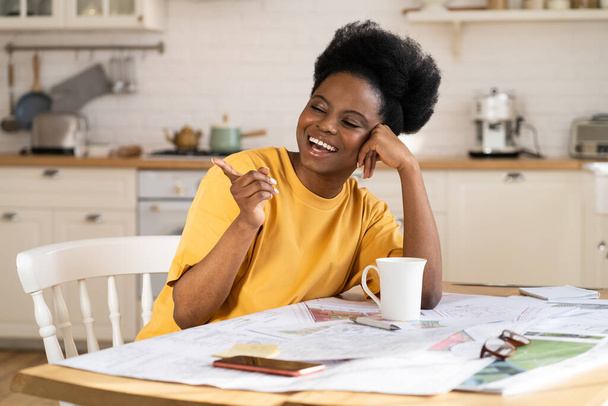 Сверхрадостная чернокожая деловая женщина смеется над работой из дома. Веселый архитектор сидит за столом с чертежами - Фото, изображение