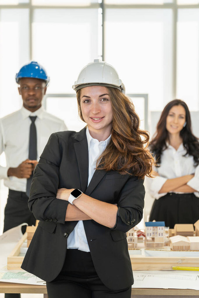 Портрет улыбающейся бизнес-леди-инженера в шлеме и коллеге по команде по многообразию с архитектурной моделью строительства. - Фото, изображение
