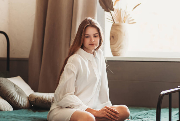 Reggeli portré egy komor fiatal lányról, hosszú hajjal egy ágyban a hálószobában. - Fotó, kép