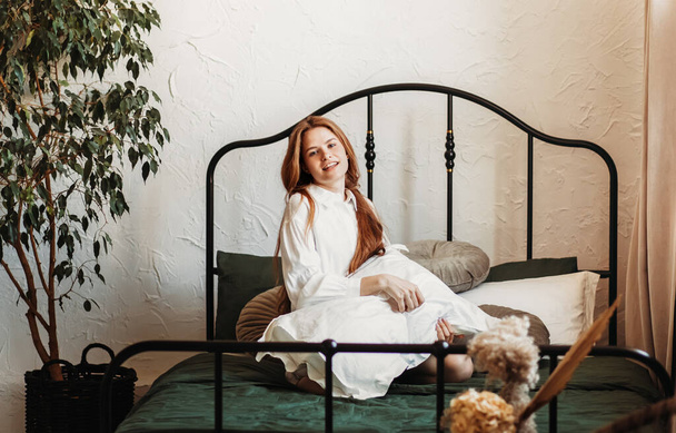 Young happy smiling girl with long hair sits with a pillow on the bed in the bedroom - Photo, Image
