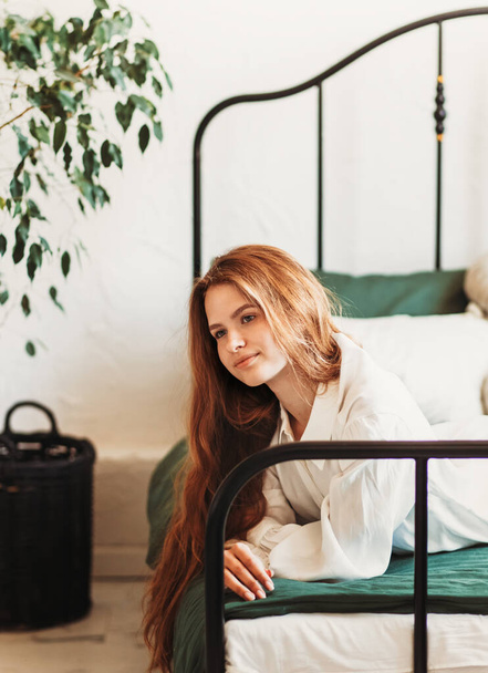 Retrato de una hermosa chica con el pelo largo en ropa blanca en el dormitorio. Una joven descansa en una cama - Foto, Imagen
