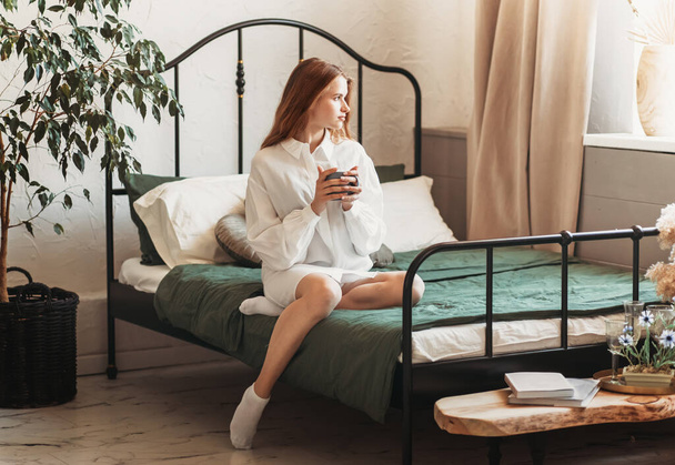 Young beautiful girl sitting on the bed with a cup of coffee in her hands - Valokuva, kuva