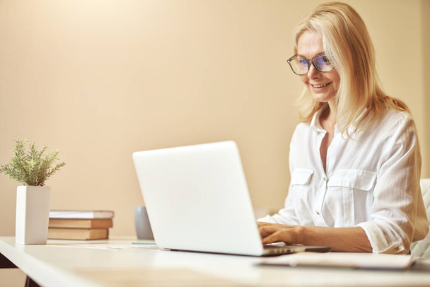 Щаслива зріла бізнес-леді в окулярах посміхається під час використання ноутбука, працює в офісі
 - Фото, зображення