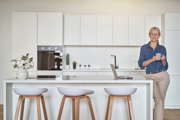 Content mature businesswoman in glasses smiling at camera, holding a cup of coffee and using laptop, standing in modern kitchen - Φωτογραφία, εικόνα