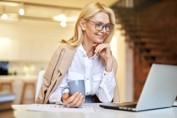 Улыбающаяся взрослая деловая женщина в очках, использующая ноутбук и пьющая кофе во время бумажной работы в офисе - Фото, изображение