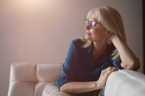 Calm mature woman in eyewear relaxing on a couch at home in the living room - Fotoğraf, Görsel