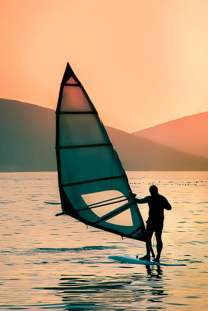 海の湾で日没の間の夕方のサーファーの自然なシルエットは、サーファーはボード上のバランスを保ち、帆、垂直、コピースペースを制御します - 写真・画像