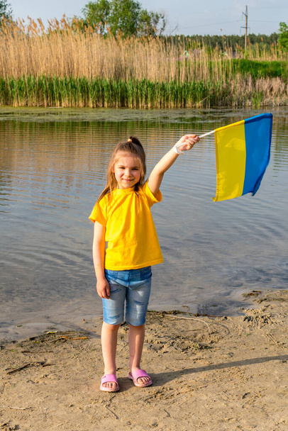 Die Flagge der Ukraine weht in den Händen eines kleinen ukrainischen Mädchens vor dem Hintergrund eines Sees und Schilfs. Unabhängigkeitstag der Ukraine und Tag der Flagge. - Foto, Bild