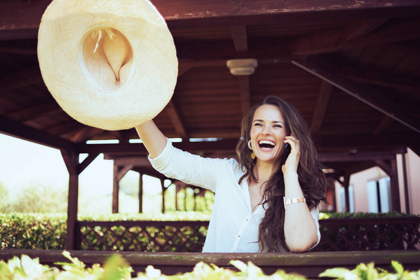 smiling trendy female in white shirt with hat talking on a smartphone on the ranch. - Photo, Image
