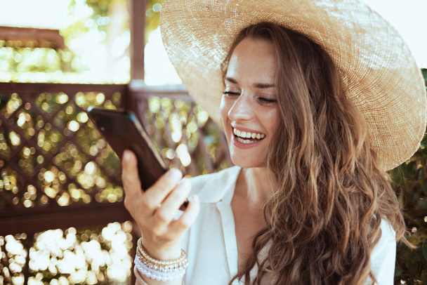 счастливая элегантная женщина средних лет в белой рубашке в шляпе с видеозвонком на смартфон на террасе гостевого дома отеля. - Фото, изображение